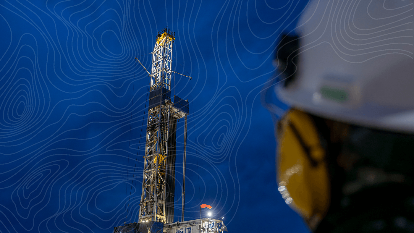 A drilling rig lit against a deep blue evening sky, viewed past the shoulder of a worker in a white hard hat and ear protection.