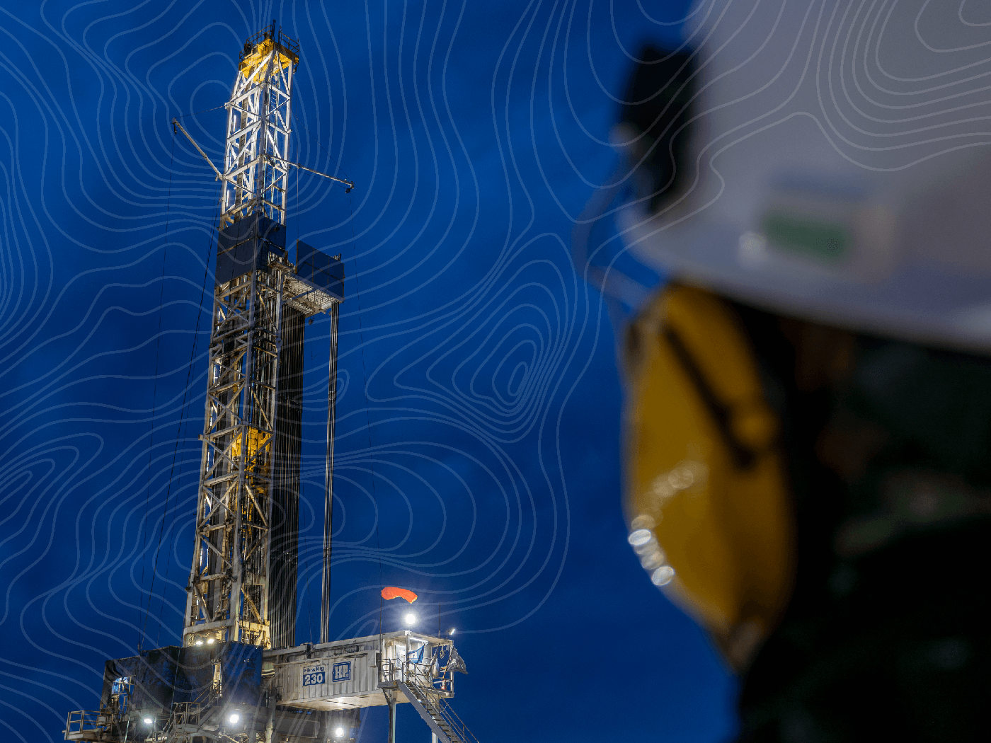 A drilling rig lit against a deep blue evening sky, viewed past the shoulder of a worker in a white hard hat and ear protection.
