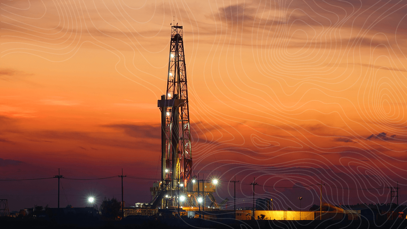 A drilling rig lit up at dusk, silhouetted against an orange sunset with power lines and site buildings along the horizon.