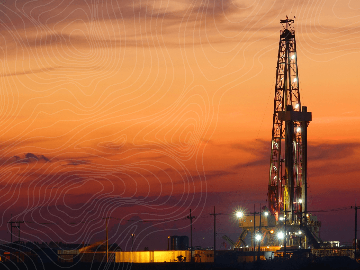 A drilling rig lit up at dusk, silhouetted against an orange sunset with power lines and site buildings along the horizon.